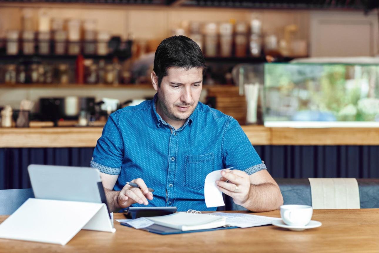 Man in a blue shirt sits at a cafe table with a tablet, taking notes and holding a receipt over papers and a notebook with a coffee cup nearby