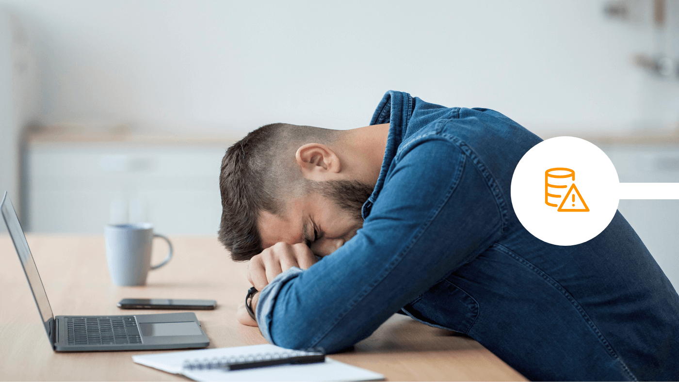 Tired man rests his head on folded arms at a desk with a laptop, notebook, phone, and mug nearby.