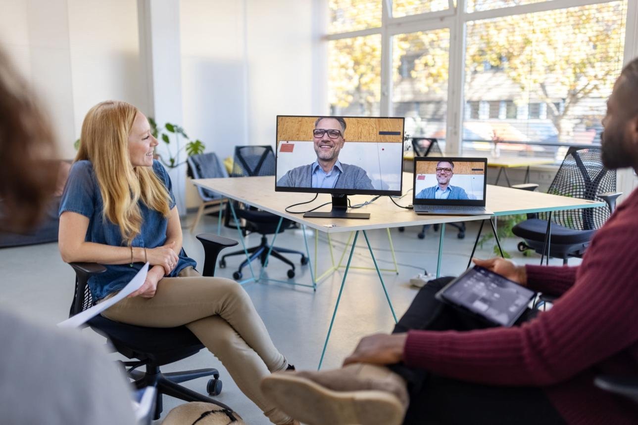 Bright office meeting with a video call: a woman on the left talks while a man appears on a large monitor and a laptop at the center of the table.