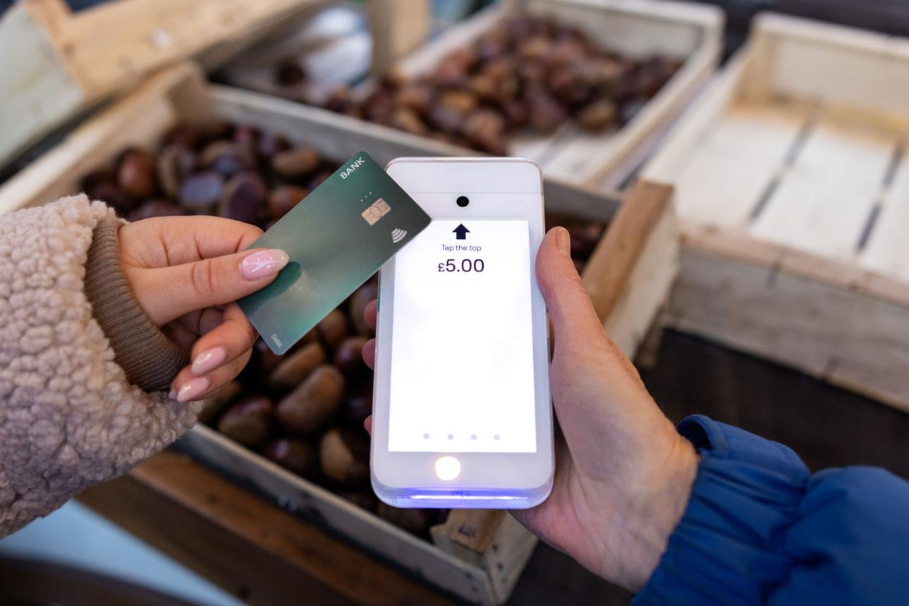 Person paying £5.00 by tapping a phone and card at a market stall with crates in the background for contactless payment.